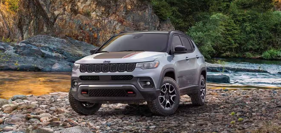 Grey Jeep Compass parked on a rocky riverbank with trees and water in the background.